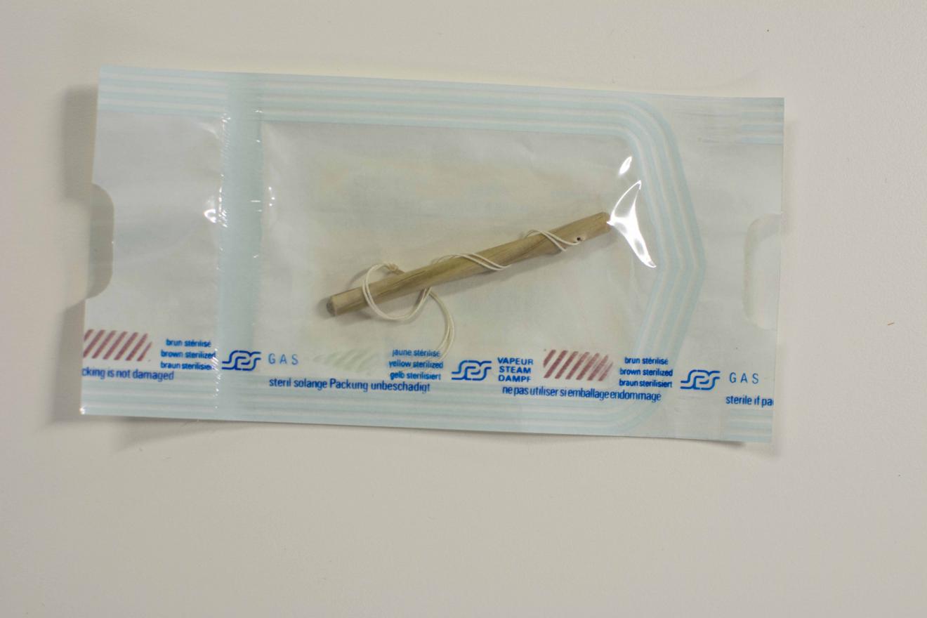 Envoltorio rectangular de papel y plástico transparente, marcado en azul y rojo con tipografía, en cuyo interior conserva una pieza tubular de material similar a la madera, en torno al que se enrolla un hilo doble de algodón