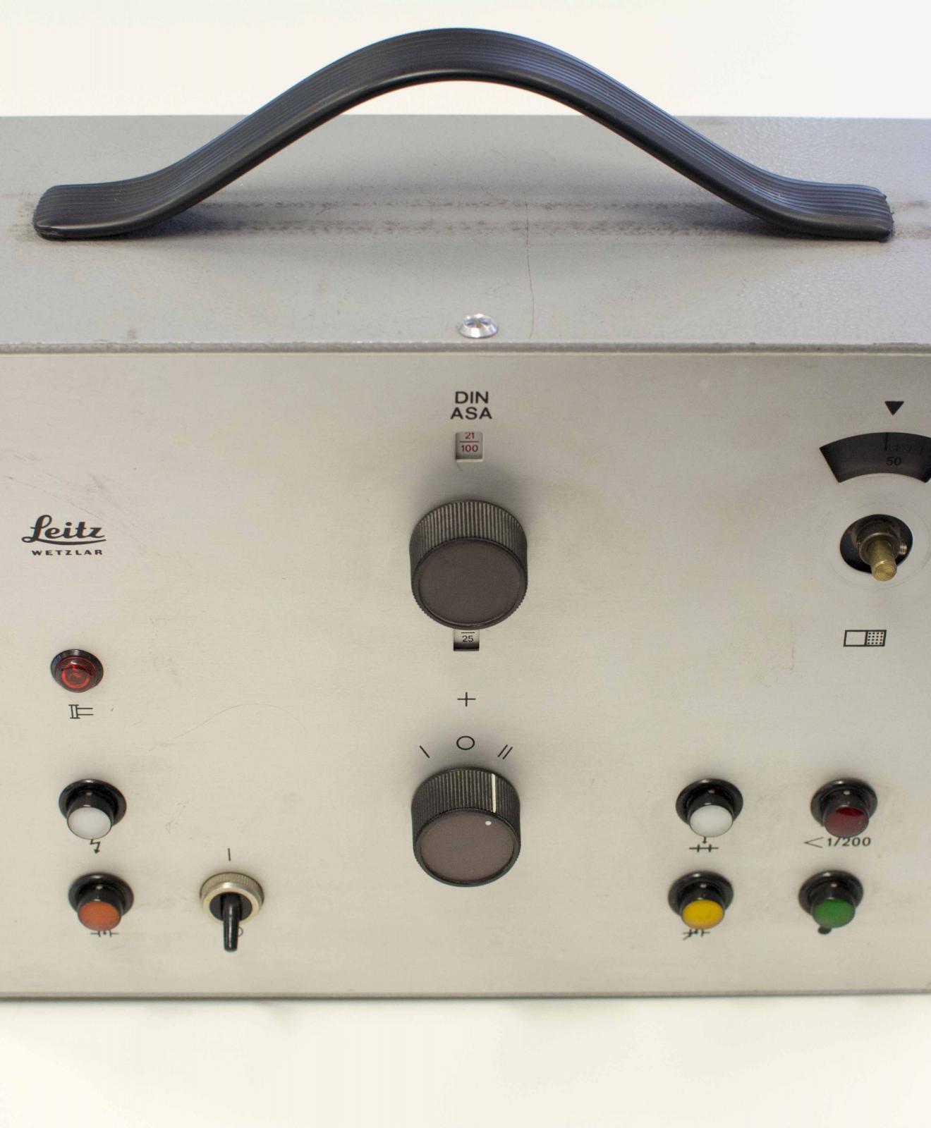 Aparato metálico con forma de maletín y asa de plástico negro. En el frontal, interruptores circulares de plástico negro con la superficie coloreada. 