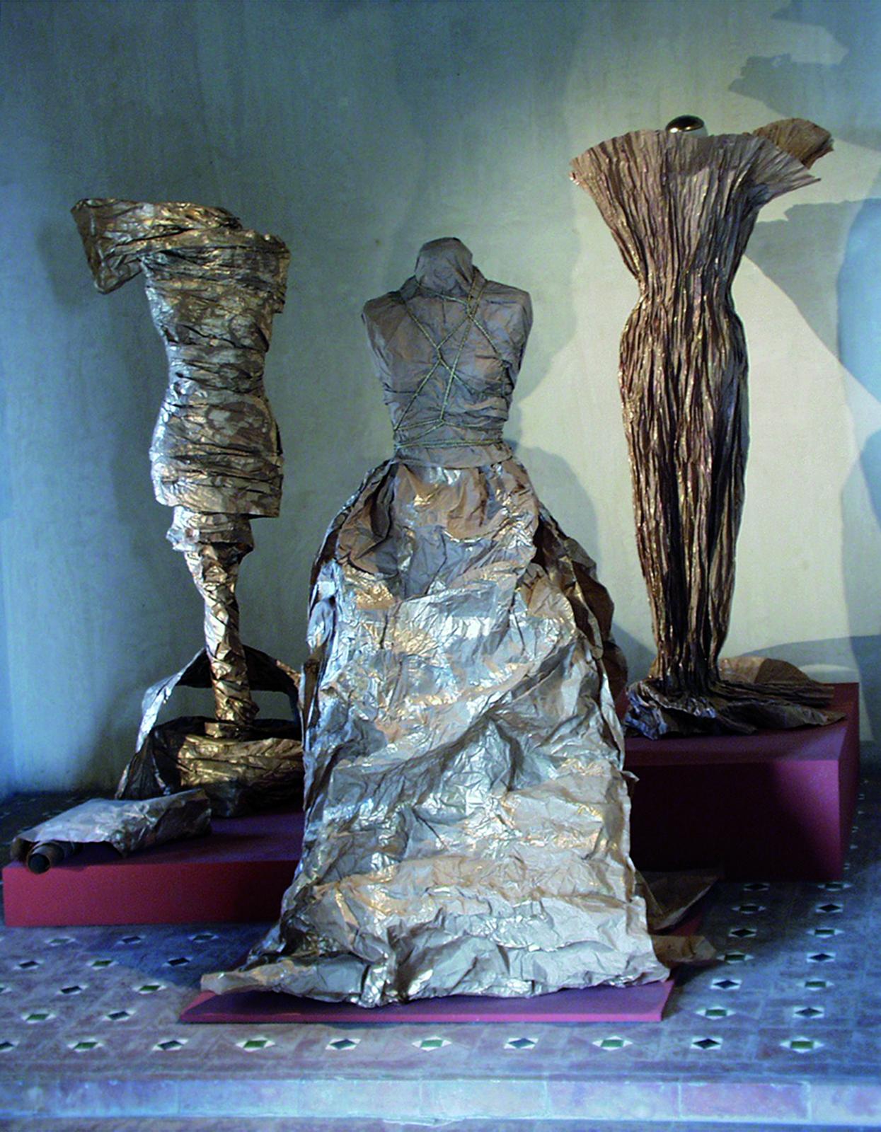 Tres vestidos de papel creados sobre maniquí y expuestos a diferentes alturas sobre peanas granates
