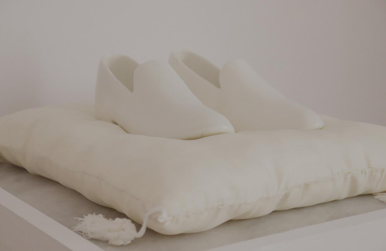 Par de mocasines blancos realizados en porcelana, sobre un cojín de seda blanco con borlones de hilo de seda colgando desde los ángulos